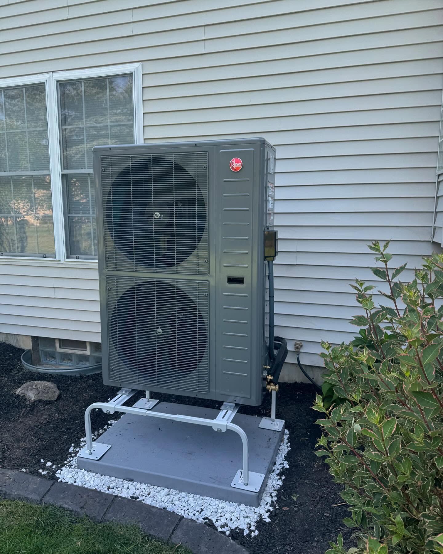 Grey heat pump unit next to a house with a window, set on a gravel base.