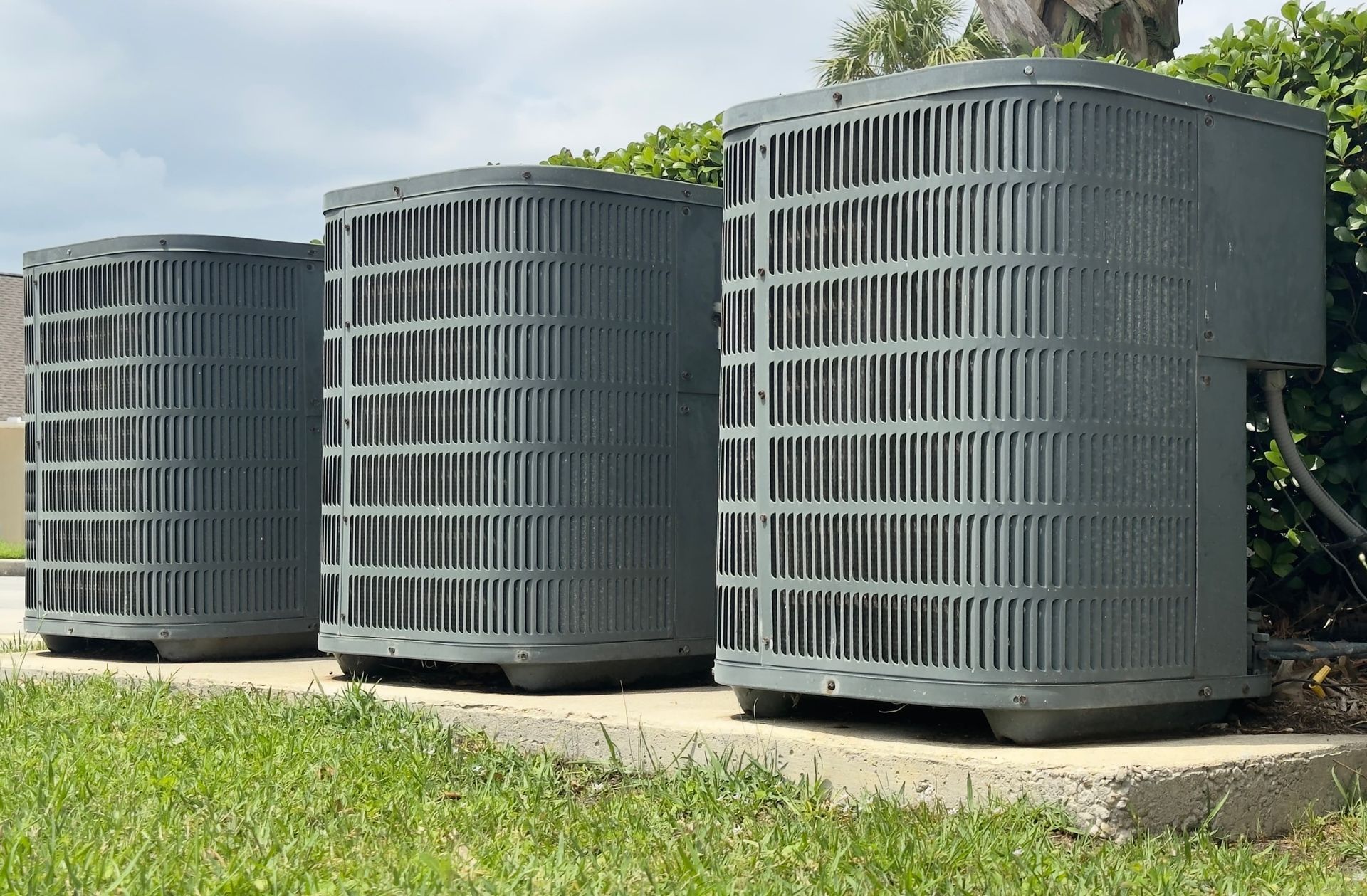 Three outdoor air conditioning units next to greenery, with dark gray metal casings.