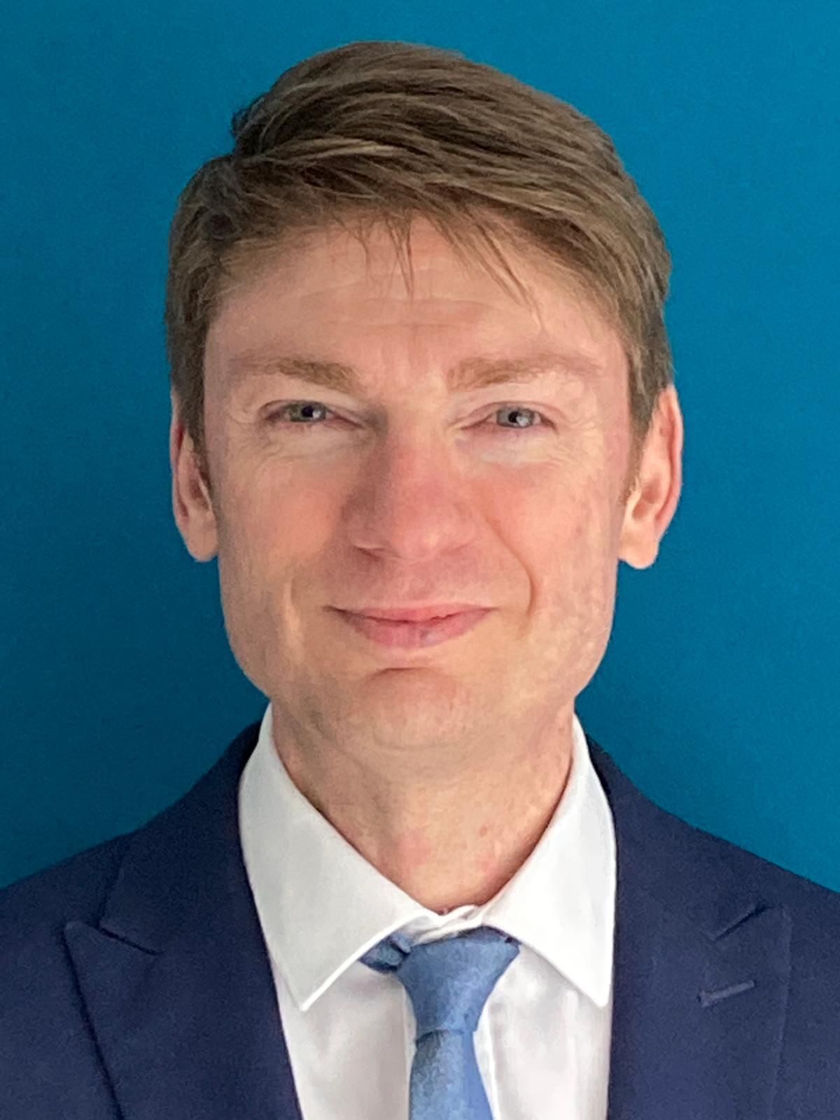 Portrait of a man in a navy suit, white shirt, and light blue tie against a blue background