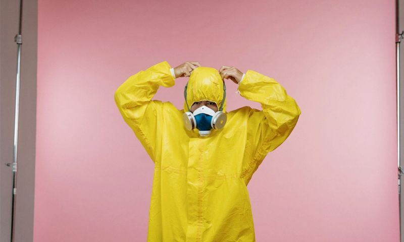 A man in a yellow protective suit and mask is standing in front of a pink background.