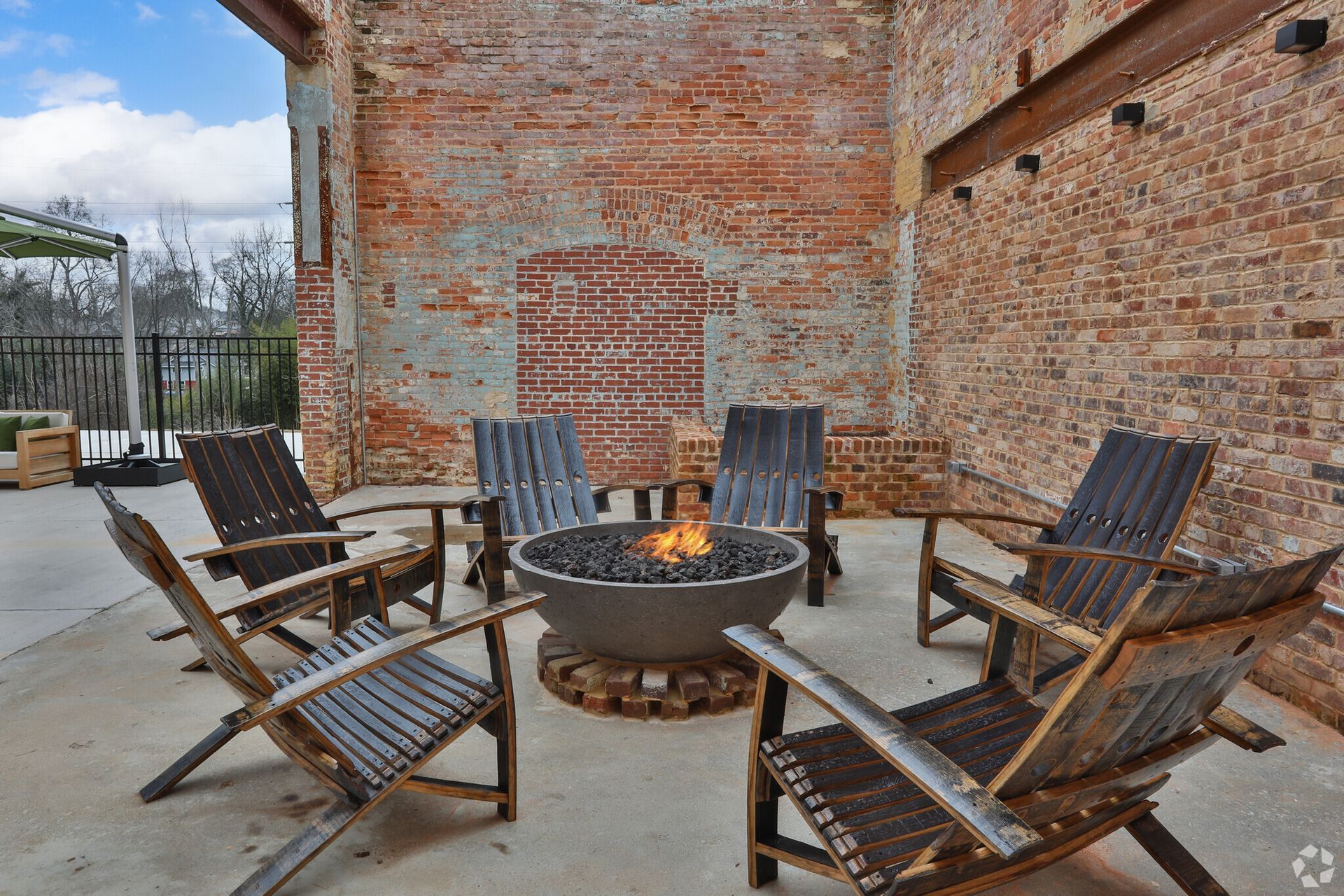 A fire pit is surrounded by wooden chairs in front of a brick wall.