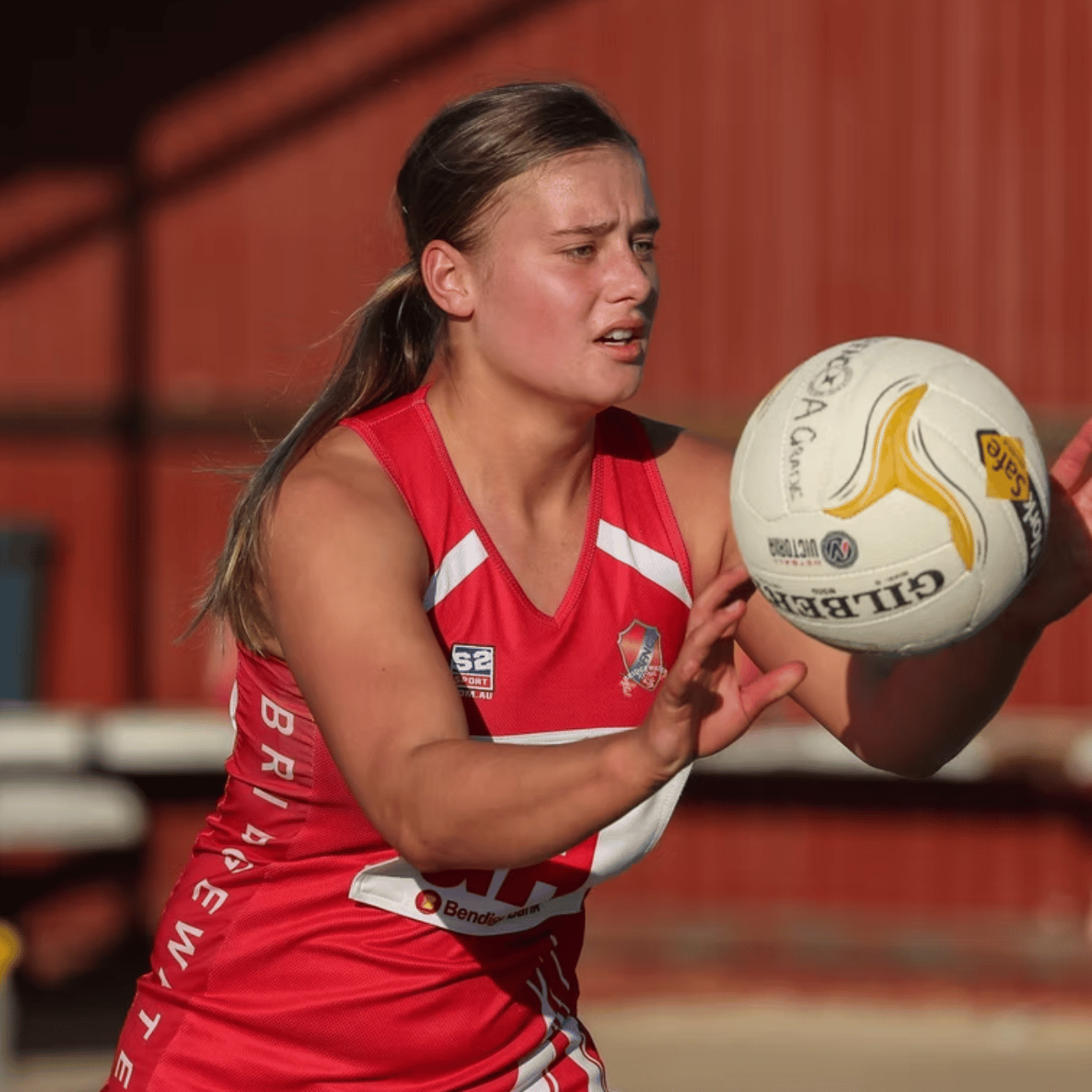 A woman in a red jersey is holding a gilbert netball