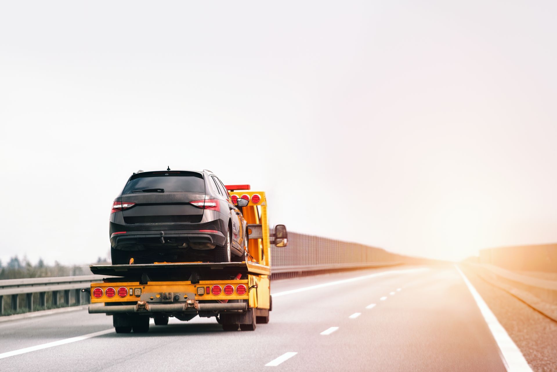 Black SUV being towed on a highway by a yellow tow truck, bright sky.