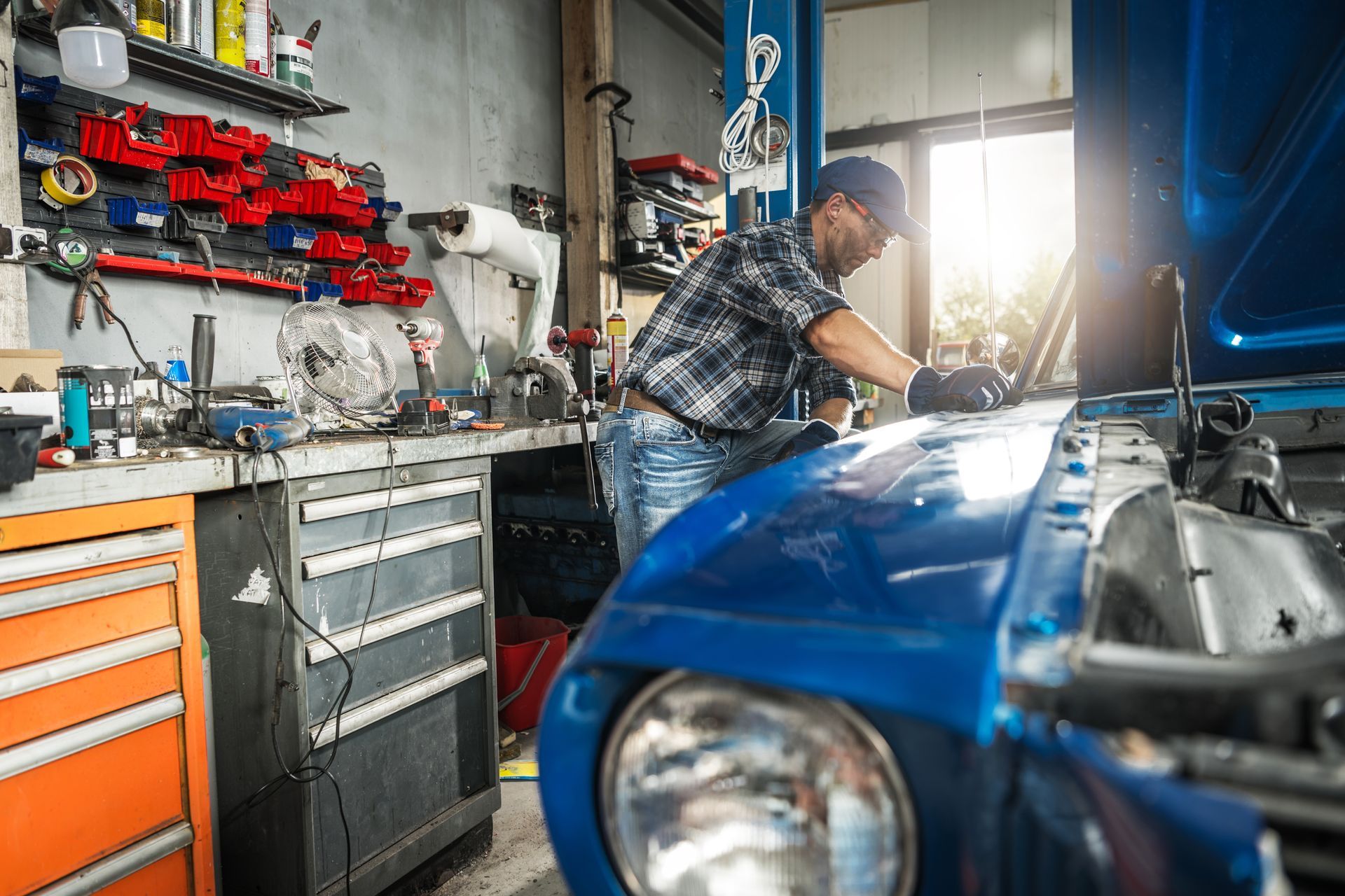 Mechanic working on a blue car in a cluttered garage, with tools and equipment.