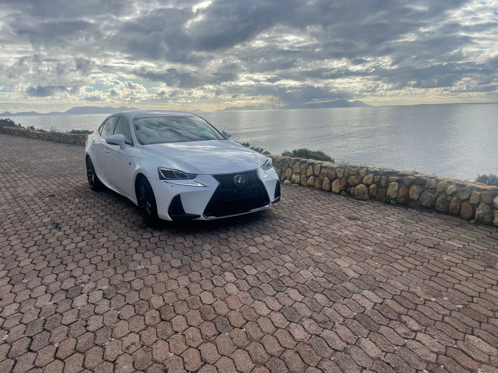 A white lexus is parked on a brick driveway next to the ocean.