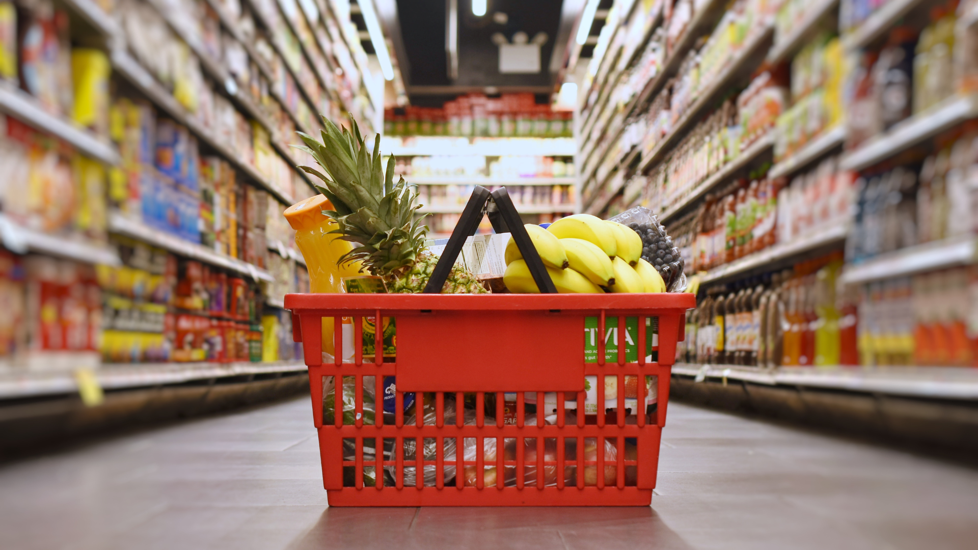 A red shopping basket filled with fruits and vegetables in a supermarket aisle.