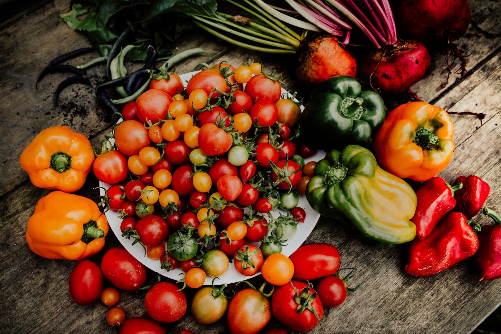 Assortment of fresh tomatoes and colorful bell peppers on a wooden surface, with beets and other vegetables.