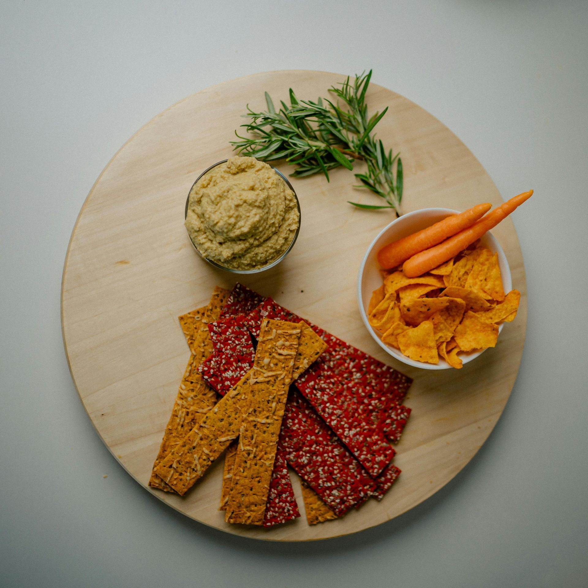 A wooden platter with hummus, crackers, carrots, and chips. Rosemary sprig on top.