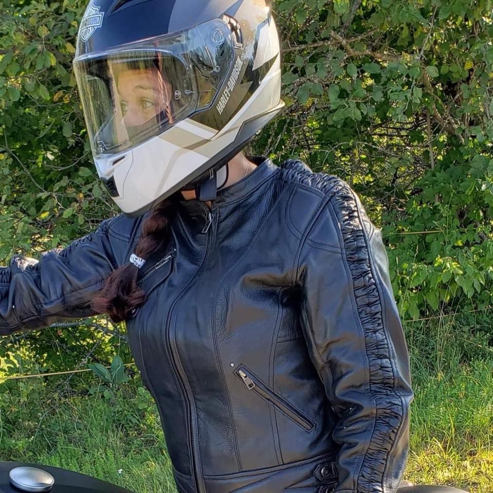 A motorcyclist wearing a dark helmet and black leather jacket sitting on a motorcycle against a background of foliage.