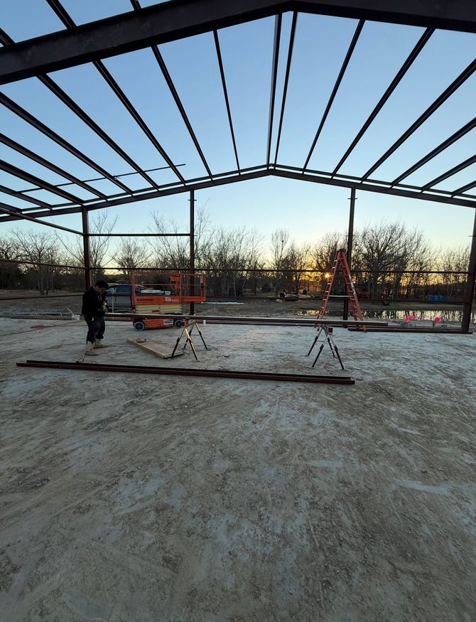 Steel building frame under construction at sunset, with workers and equipment on a gravel site.
