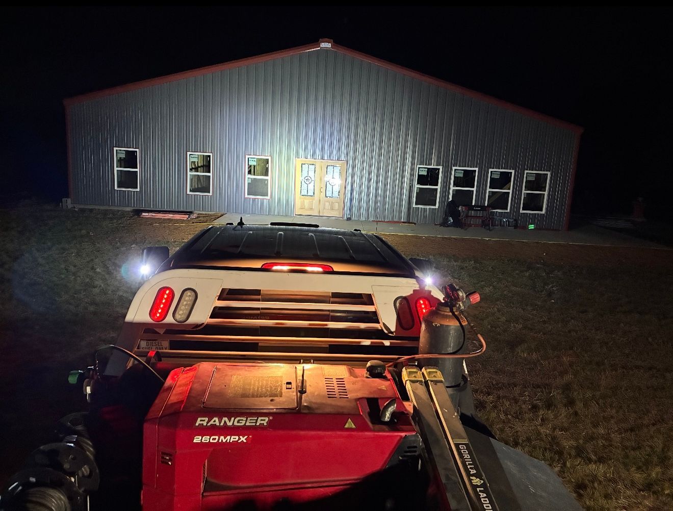 Nighttime farm scene with red tractor facing a lit metal barn, lights illuminating the building.