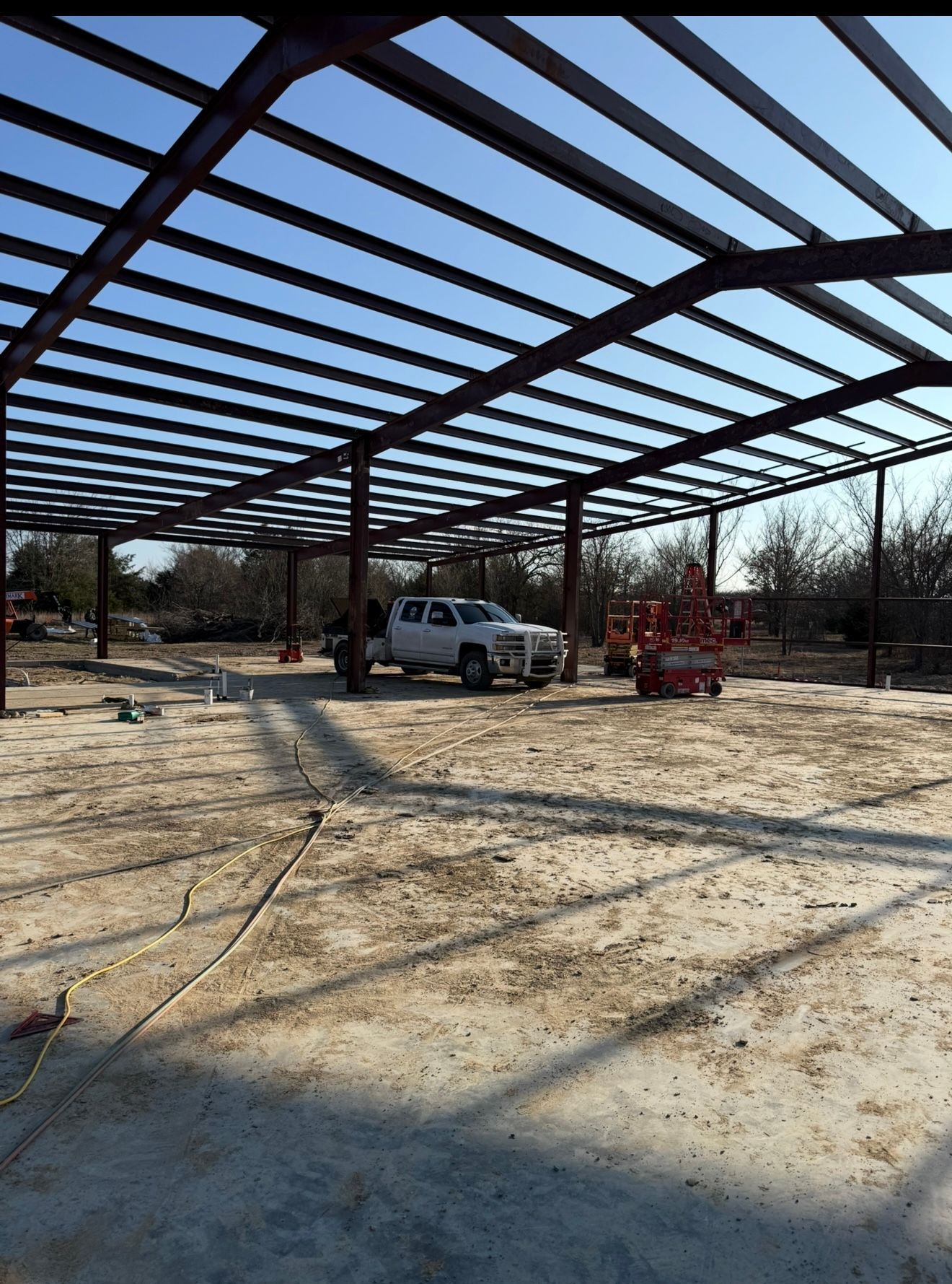 Steel building frame under construction on a gravel lot, with a white pickup truck and red machinery.