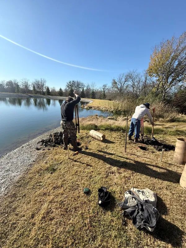 Two people working by a lakeshore on a sunny day, with gear and bags on the ground.