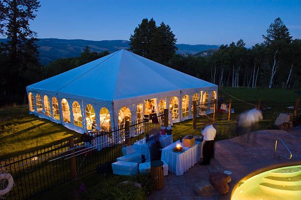 Large, illuminated white tent on a lawn at dusk; table with food and drinks, pool nearby, trees and hills in the background.