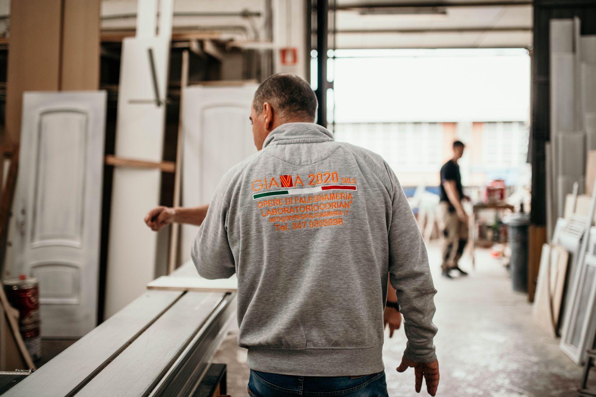 Uomo con giacca grigia in un'officina, che indica, con un logo sulla schiena; un altro uomo sullo sfondo.