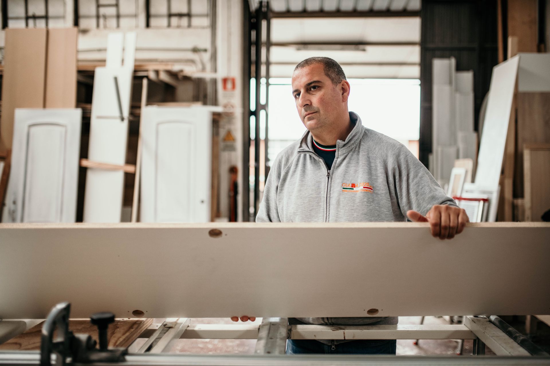 Un uomo con un maglione grigio in una falegnameria osserva attentamente una grande lavagna bianca.