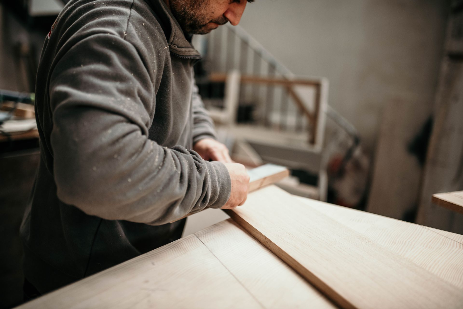 Un falegname che leviga un'asse di legno, al lavoro in un'officina, indossa una felpa grigia.