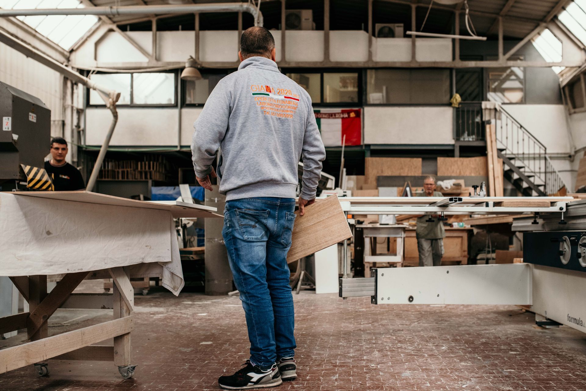 Un uomo con una felpa grigia trasporta legna in un'officina; sono visibili altri operai e attrezzature.