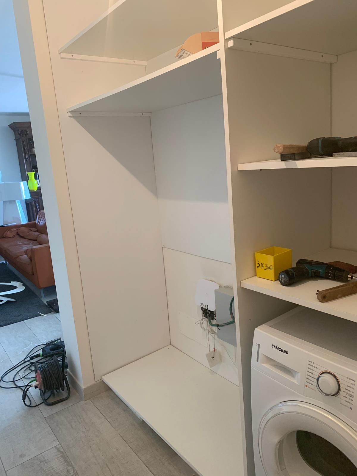 Armadio a muro bianco con lavatrice. Quadro elettrico e mensola. Pavimento in legno chiaro.