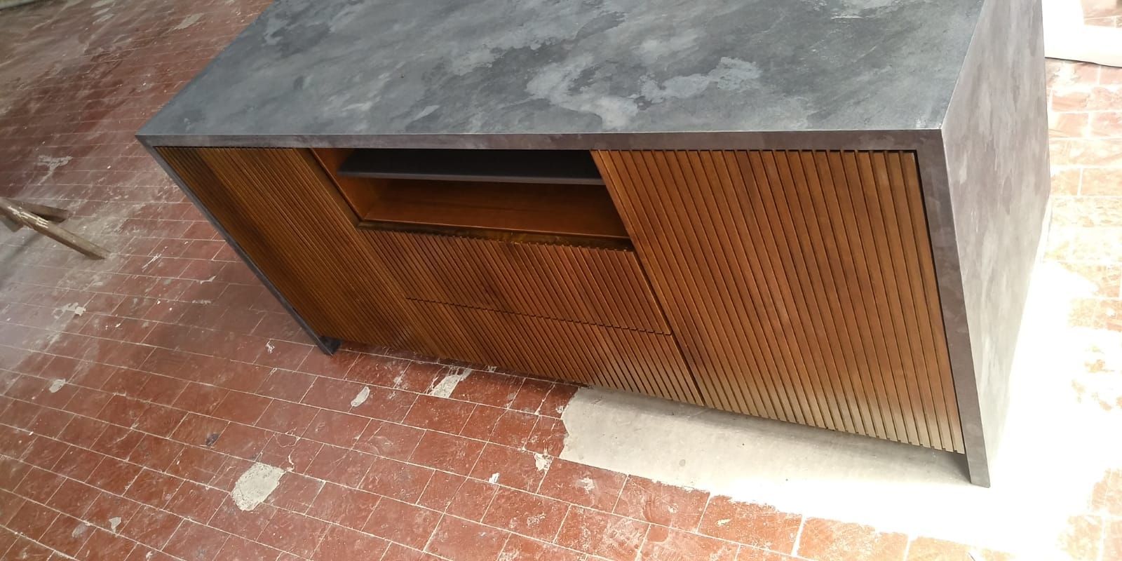 Un mobile in legno con un piano di lavoro grigio e ante traforate su un pavimento piastrellato rosso.
