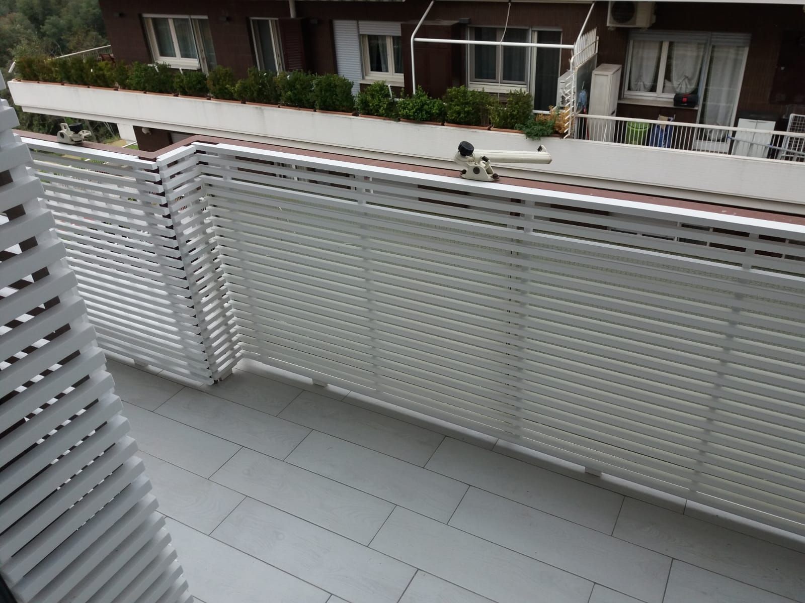 Recinzione in listelli bianchi su un balcone con pavimento in piastrelle grigie; edificio sullo sfondo.