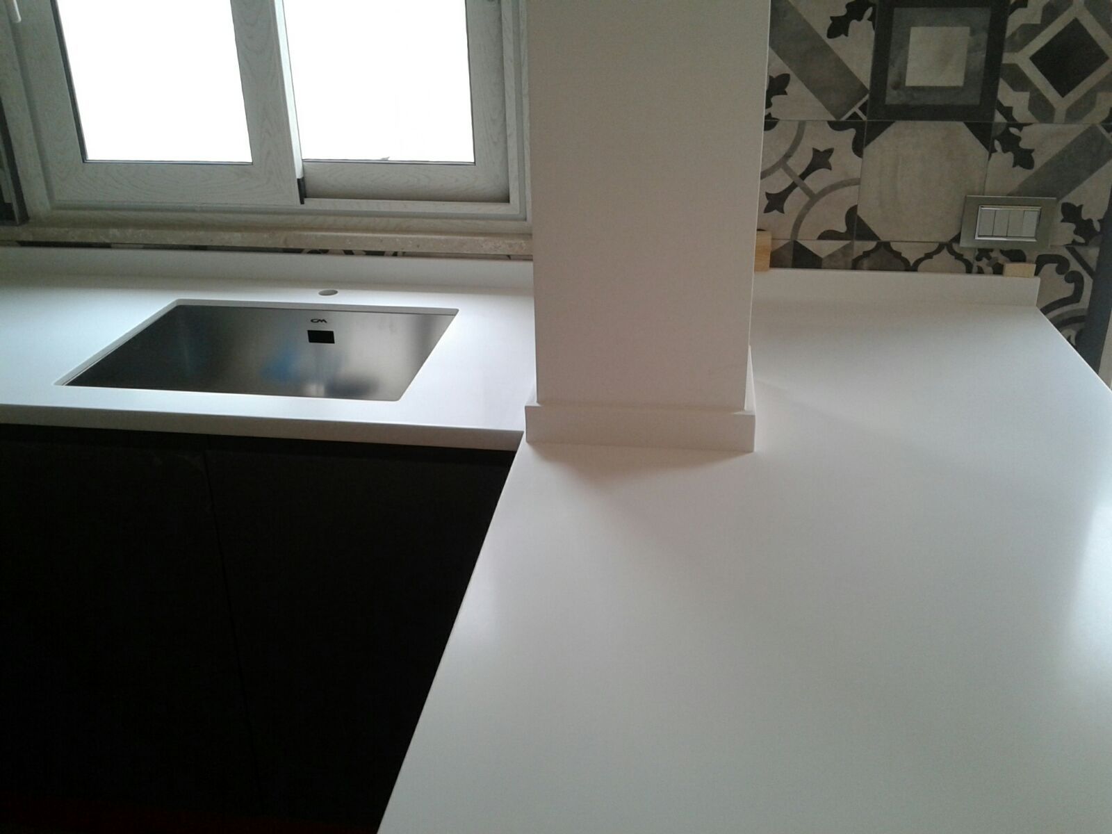 Cucina con piano di lavoro bianco, lavello in acciaio inossidabile, mobili scuri e finestra.