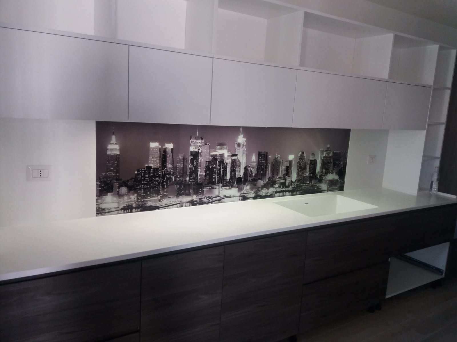 Cucina moderna con paraschizzi raffigurante un paesaggio urbano in bianco e nero, ripiani bianchi e mobili scuri.