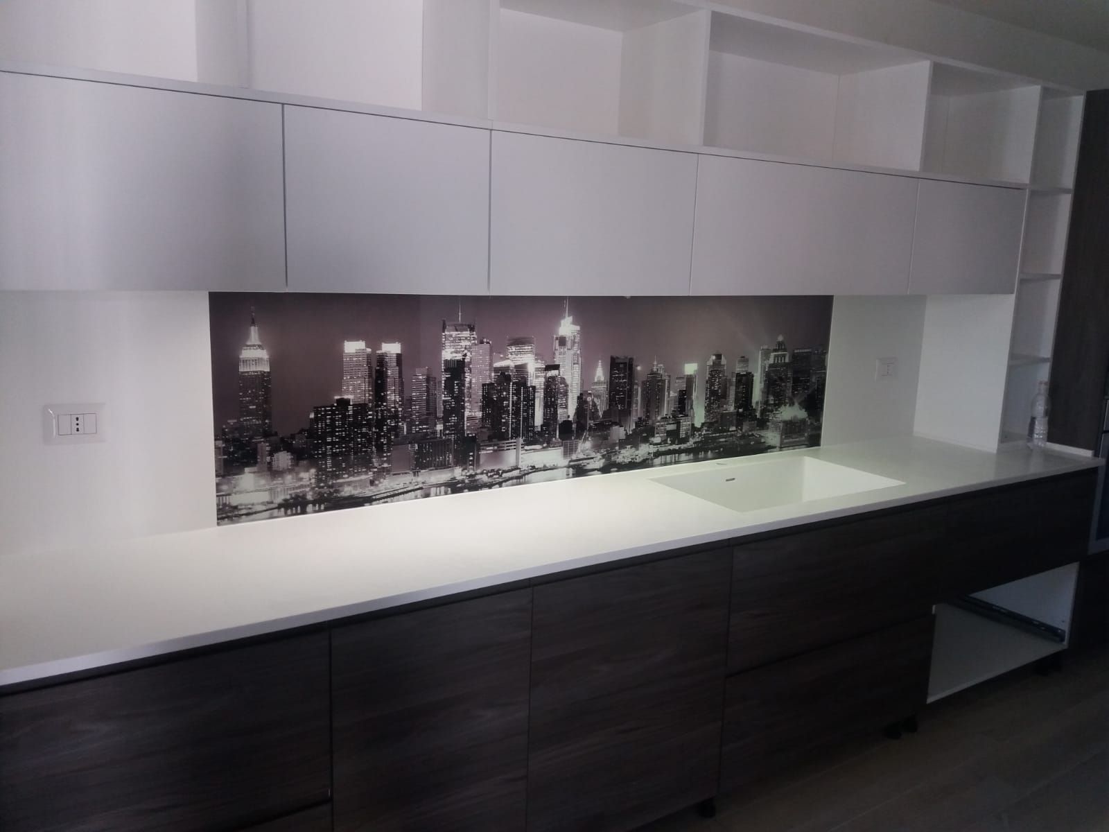 Cucina con mobili scuri, ripiani bianchi e paraschizzi con motivi urbani. Sopra ci sono mobili contenitori bianchi.