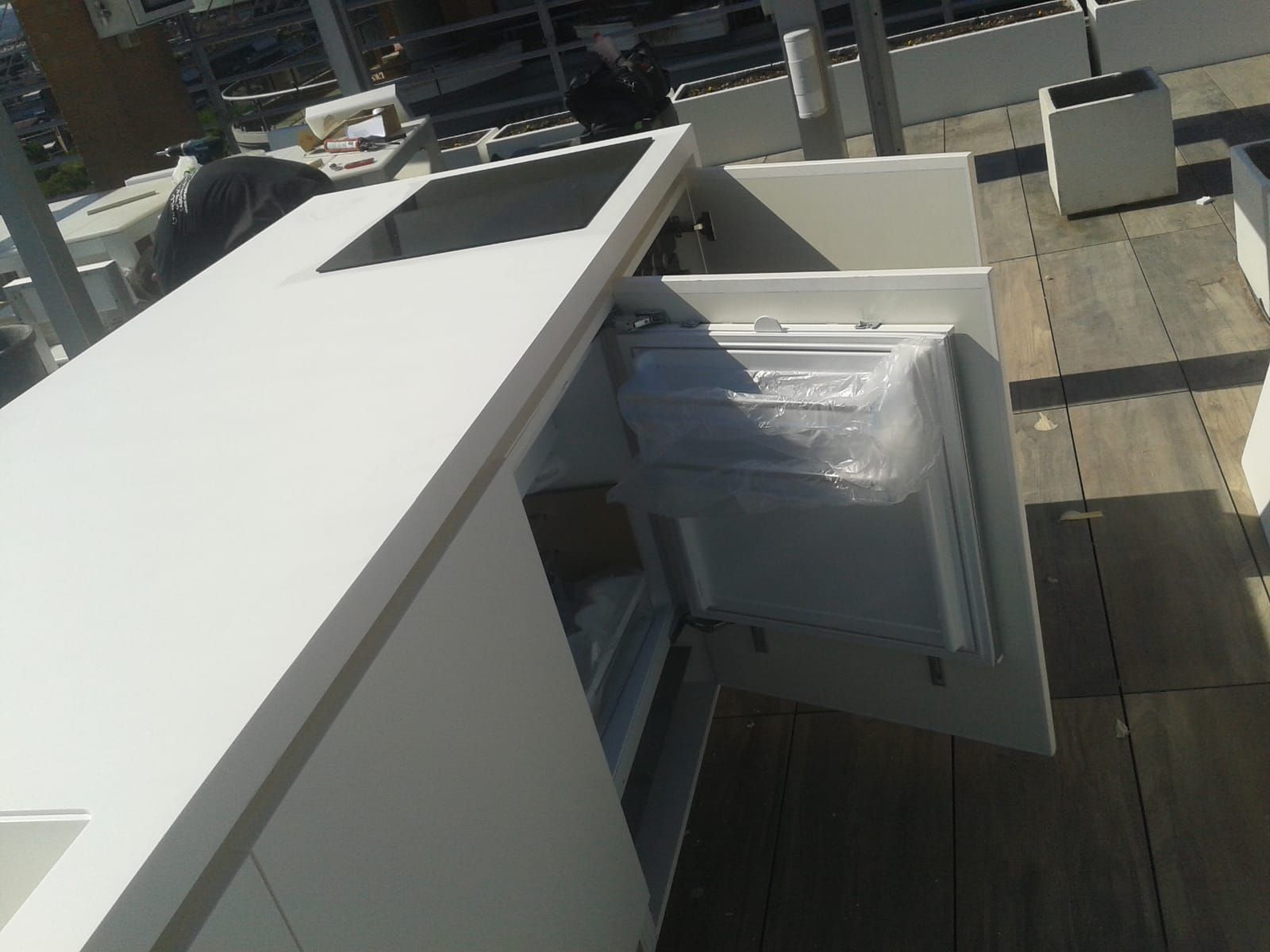 Cucina bianca da esterno con cassetto del frigorifero aperto su una terrazza in legno, soleggiata.