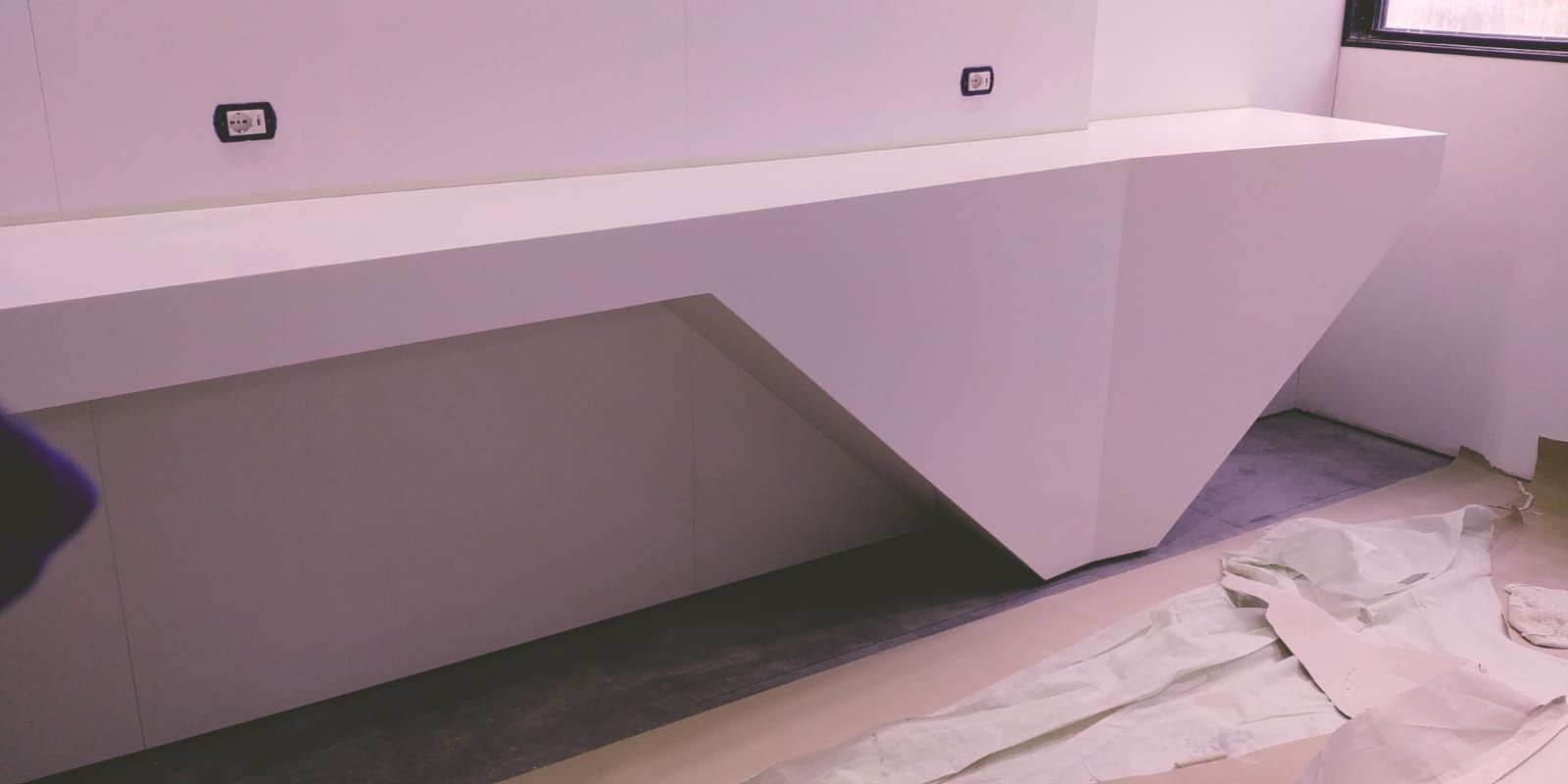 Una scrivania bianca, angolare, incassata, con una netta forma a V rovesciata. Le prese elettriche sono visibili.