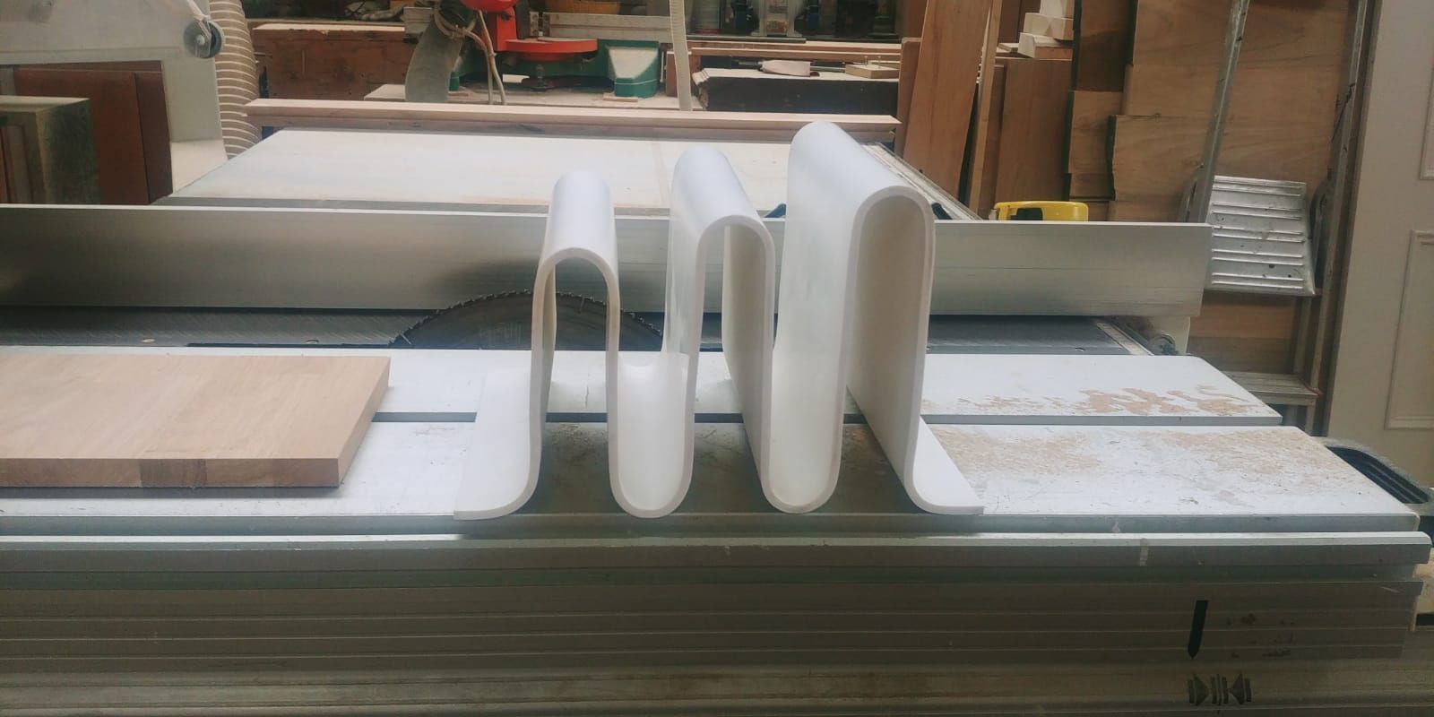 Oggetti bianchi curvi su una sega circolare. Un blocco di legno sulla sinistra. Falegnameria sullo sfondo.