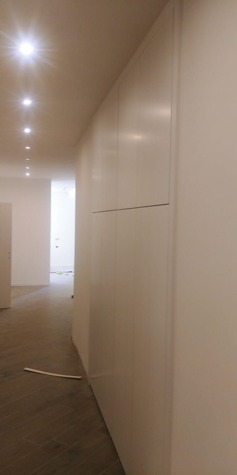 Un corridoio lungo e stretto con pareti bianche e un pannello crepato; luci da incasso brillano dal soffitto.
