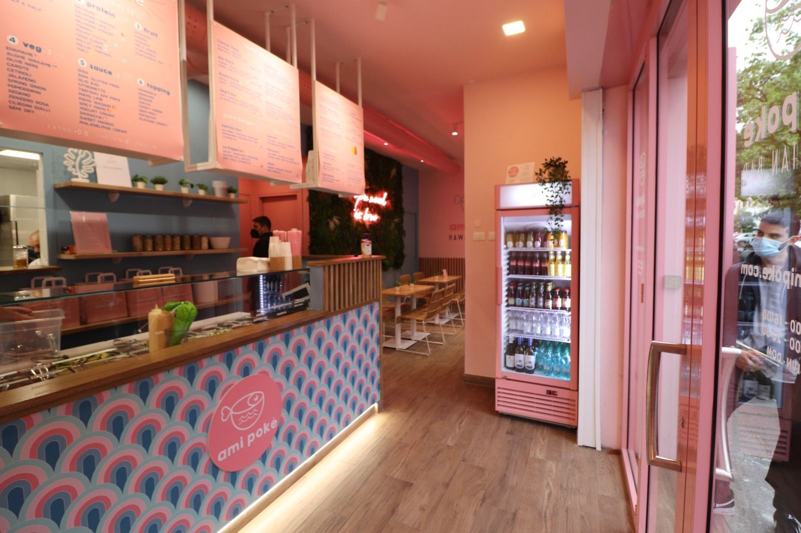 Interno di un bar rosa con menù, bancone con design geometrico e frigorifero per le bevande.