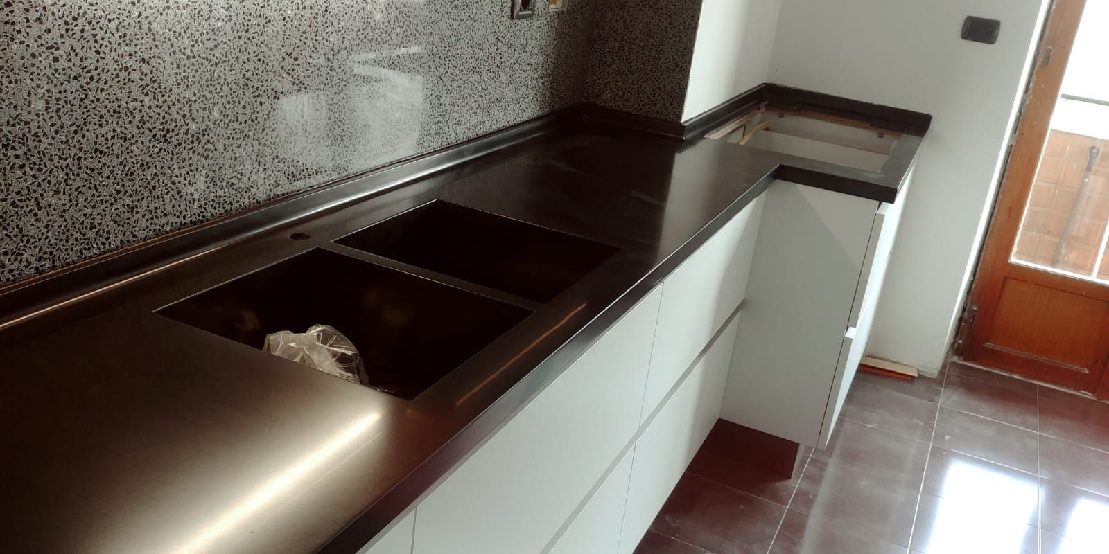 Piani di lavoro scuri della cucina con doppio lavello e paraschizzi maculato. Mobili bianchi e porta in legno.