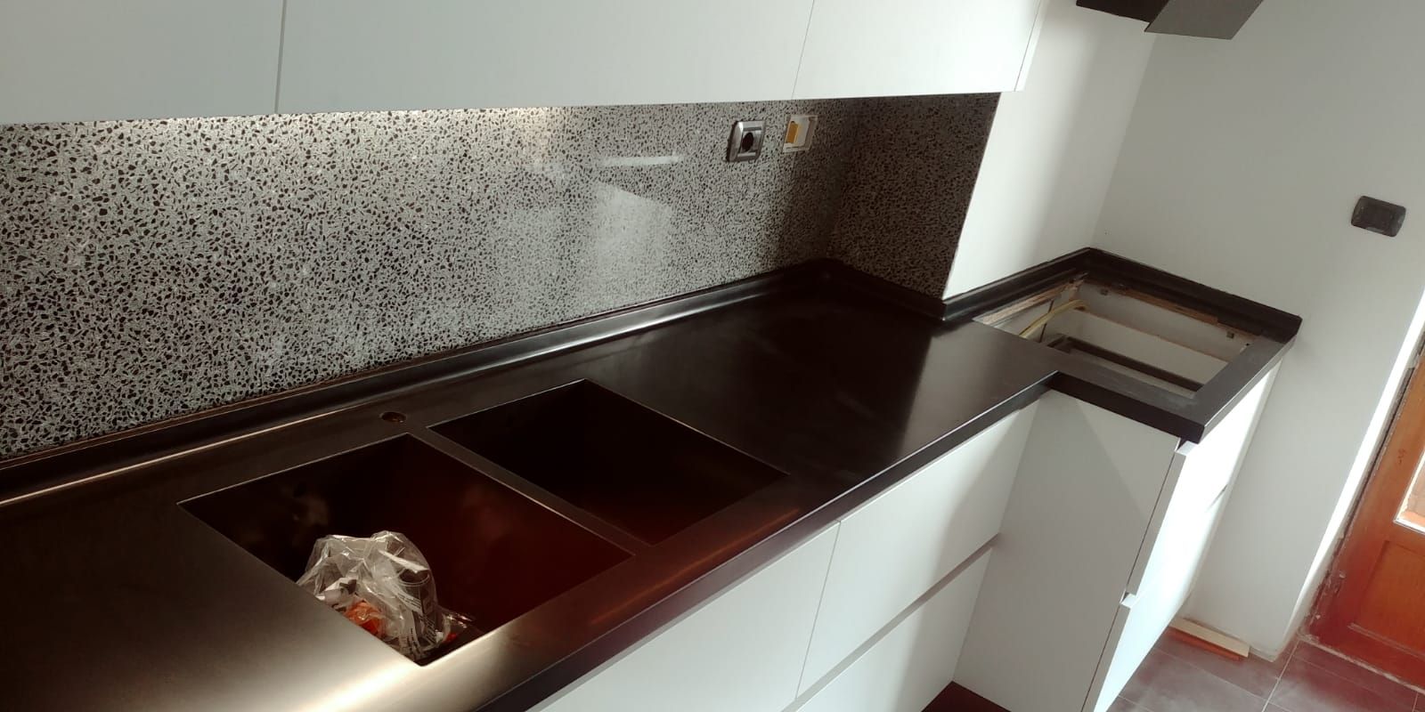 Una cucina moderna con piano di lavoro scuro e un grande lavello. Mobili bianchi e paraschizzi in mosaico.