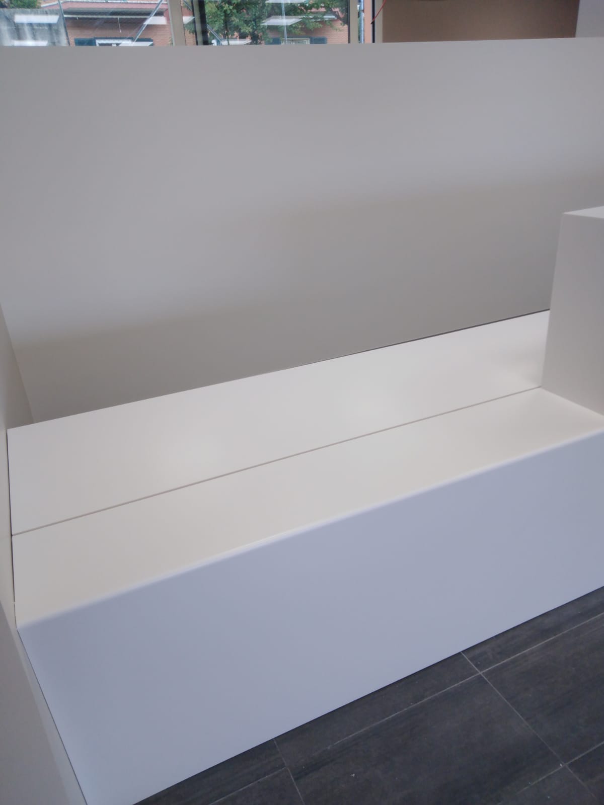 Panca rettangolare bianca contro una parete bianca con pavimento piastrellato grigio scuro.