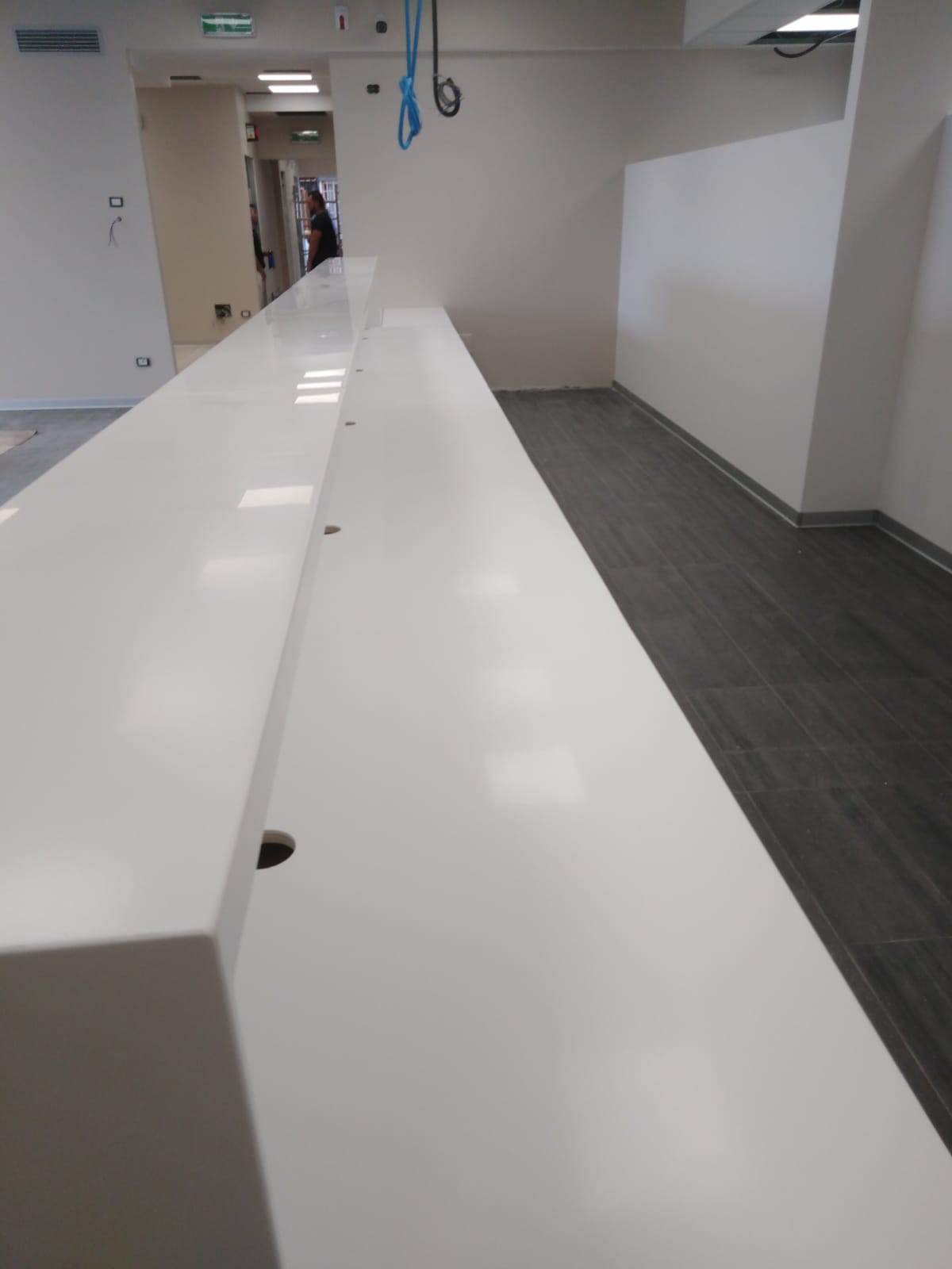Lungo bancone bianco in un moderno ufficio, con fori visibili. Pavimento scuro e pareti bianche.