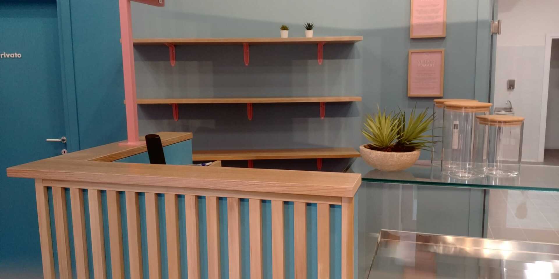 Interno di un bar con bancone in legno, scaffali e piante decorative. Pareti turchesi e rosa.