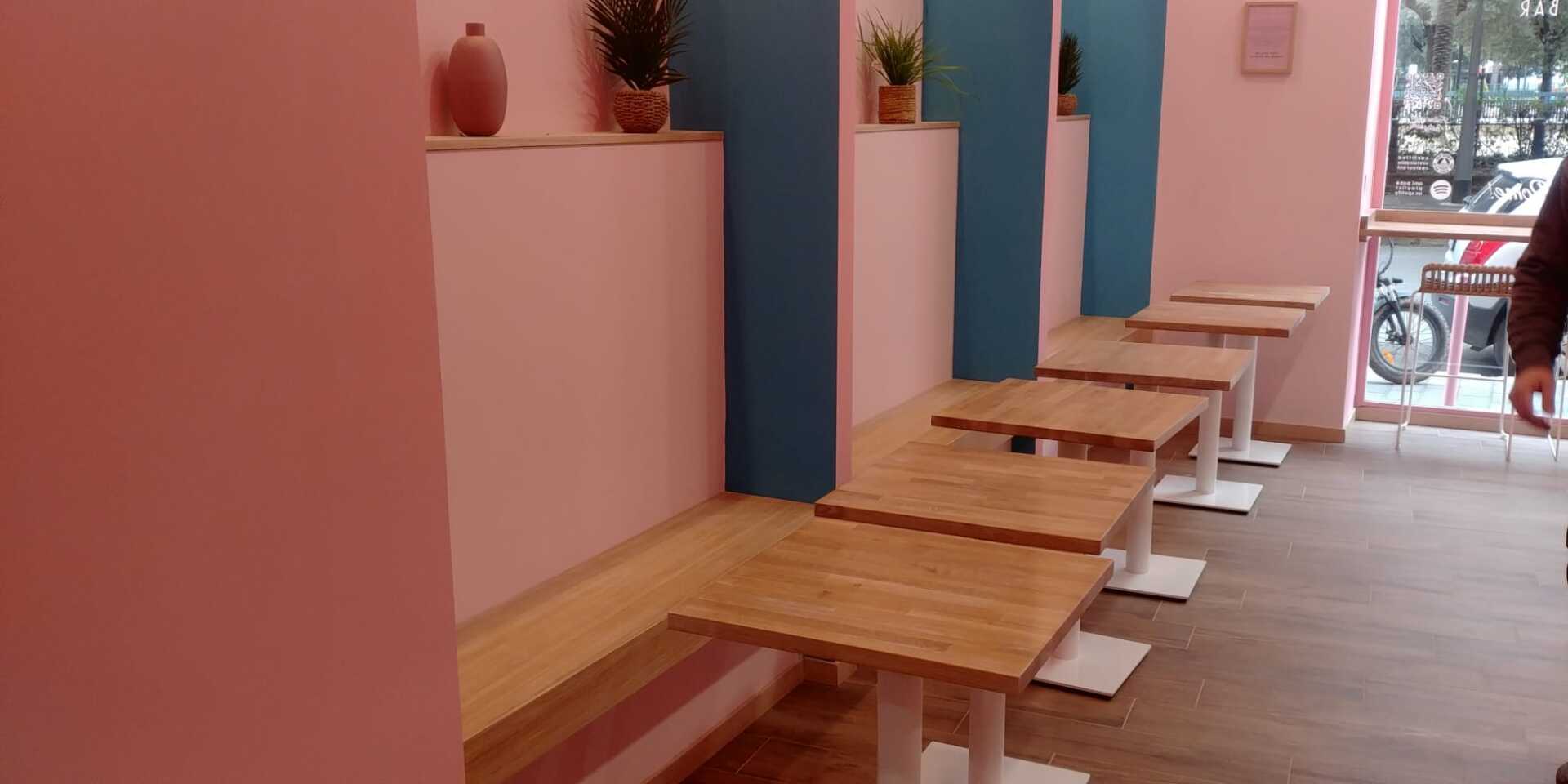 Interno di un bar con pareti rosa, tavoli in legno e pilastri blu. Una persona è in piedi vicino a una finestra.