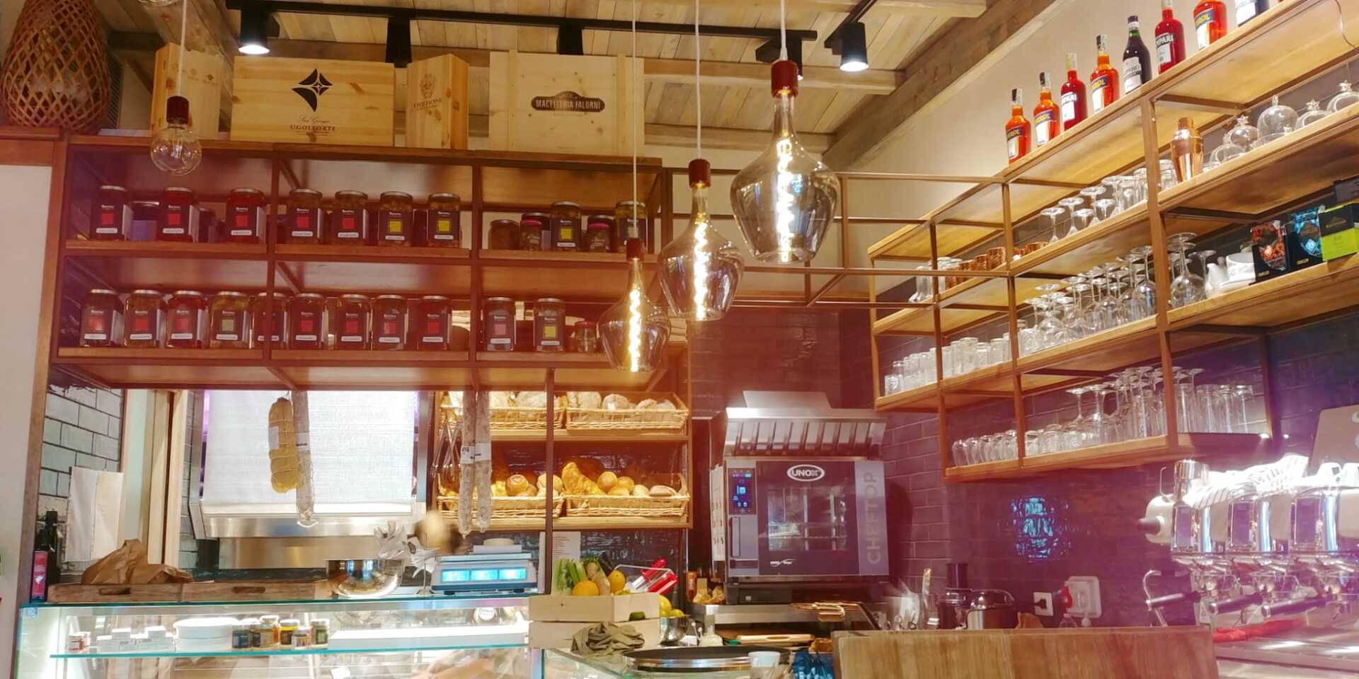 Interno di un bar con scaffali pieni di prodotti, bancone, luci sospese e una macchina per il caffè.