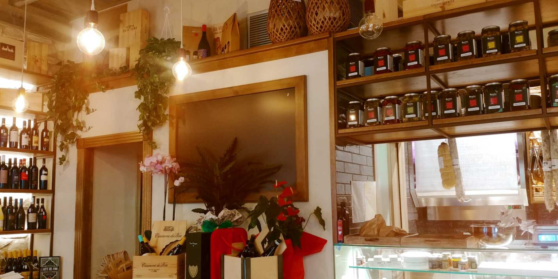 Interno di un'enoteca con scaffali pieni di bottiglie di vino, cibo e piante decorative.