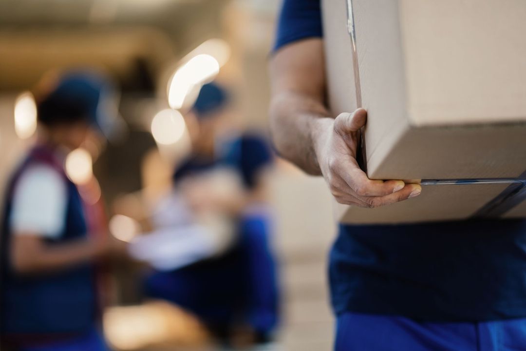 Un uomo in uniforme blu trasporta una scatola di cartone; sullo sfondo si vedono magazzinieri sfocati.