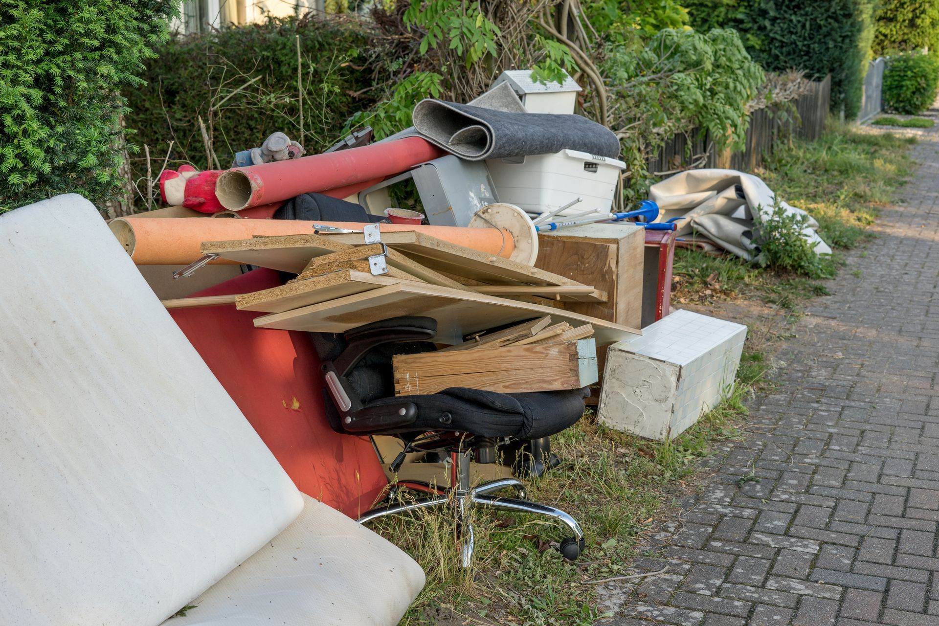 Mucchio di mobili abbandonati e detriti su un ciglio erboso accanto a un marciapiede, con una siepe sullo sfondo.