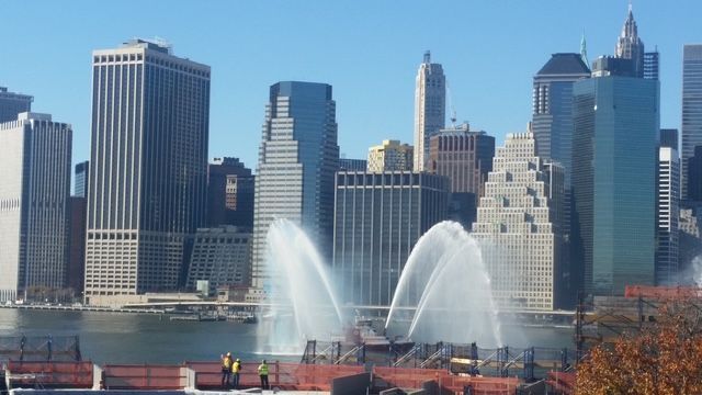 Skyline of New York City with water fountains spraying in the foreground on a sunny day.