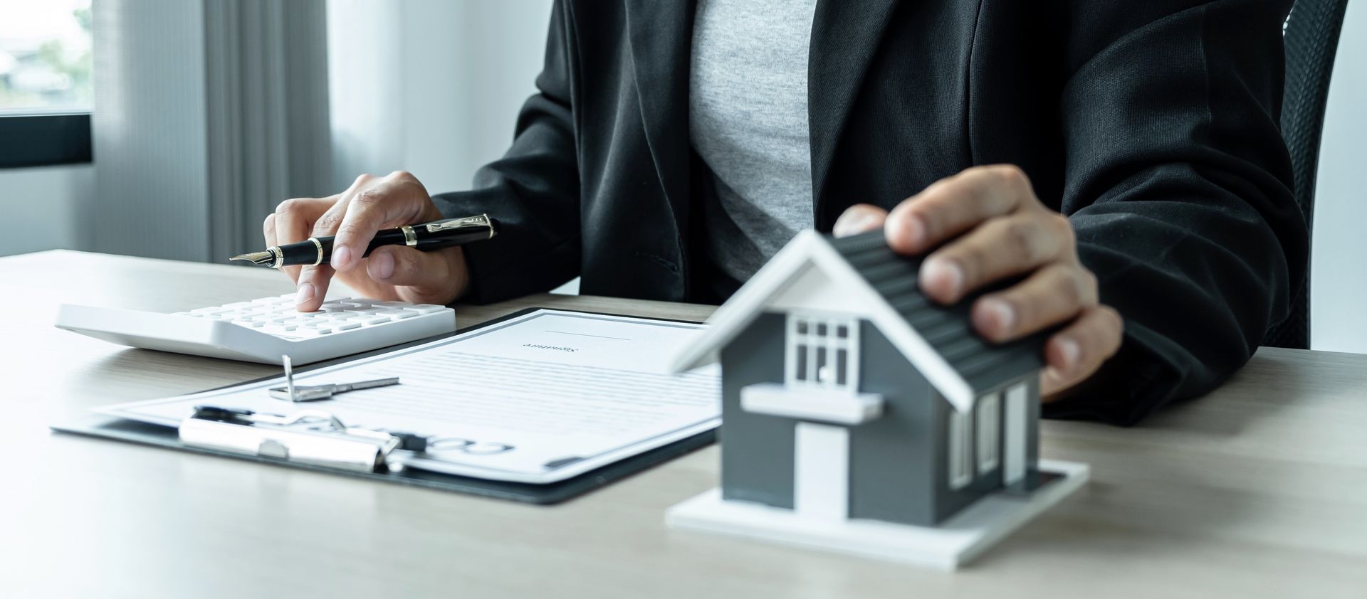 Person reviews documents and a calculator beside a small house model on an office desk.