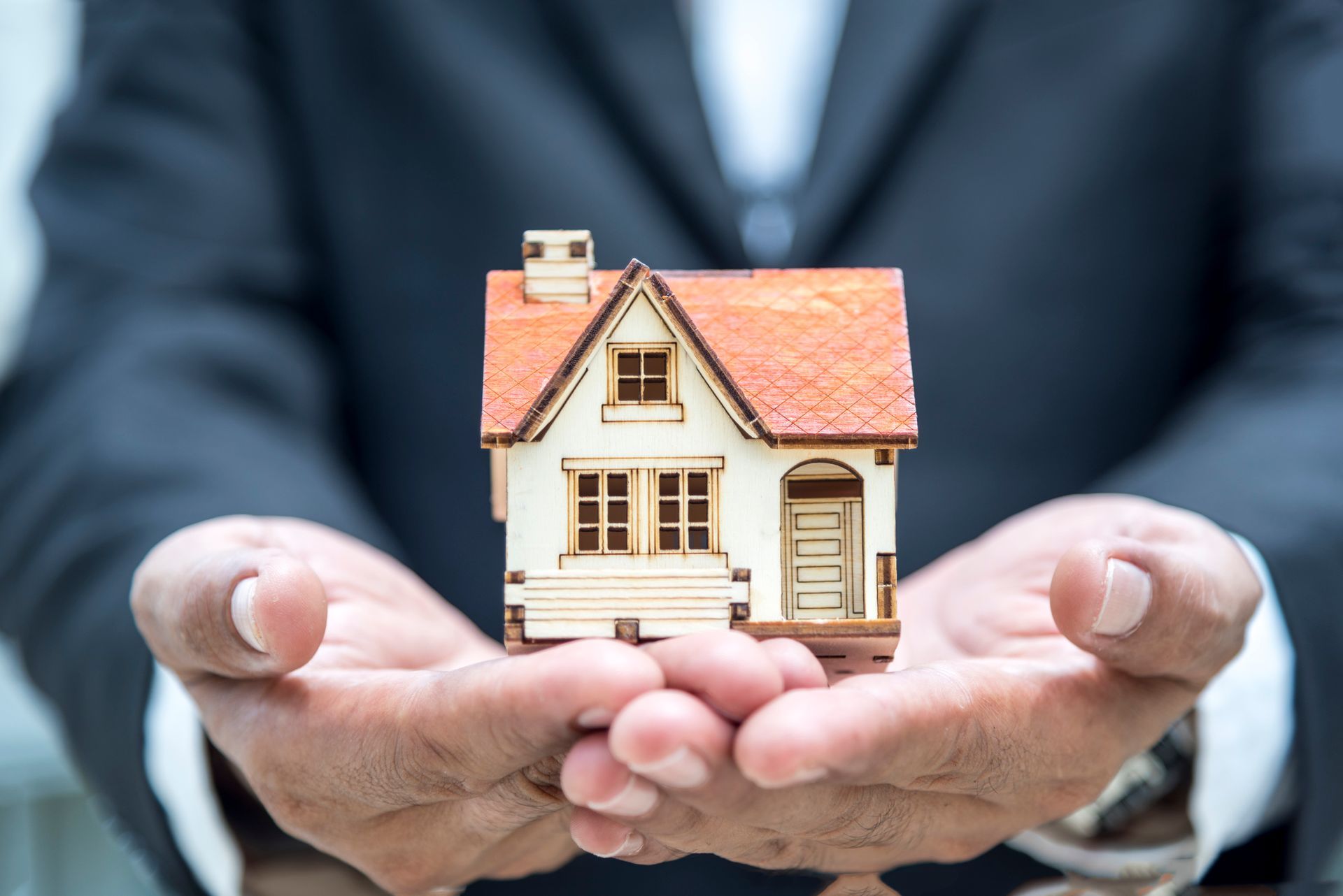 Person in a suit holding a small model house with both hands.