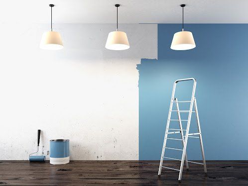Room being painted, half white, half blue, with ladder, paint can, roller, and pendant lights.