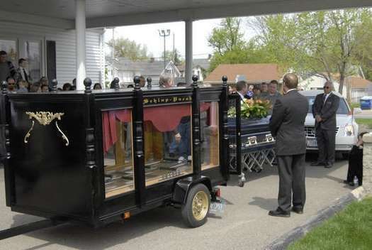 A man in a suit is standing next to a coffin on a trailer.