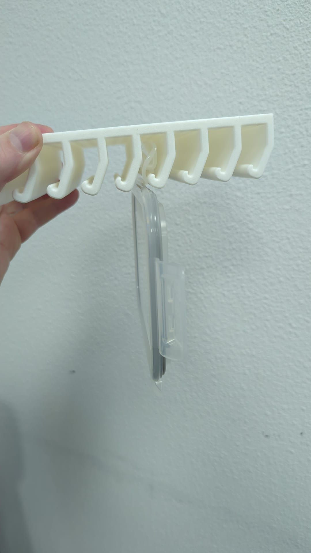 Rig-3d Een hand houdt een wit, 3D-geprint rekje met zeven haken tegen een effen muur, bevestigd met een transparante plakstrip.