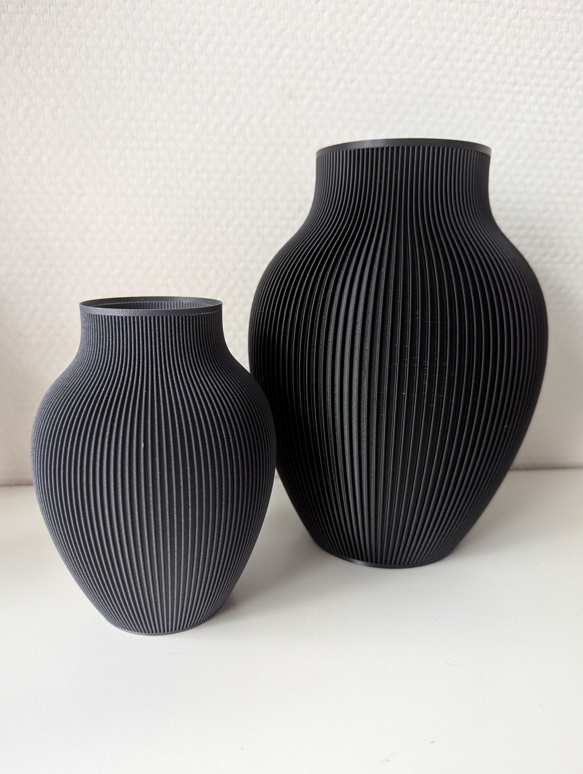 Rig-3d Twee zwarte, 3D-geprinte vazen met een gestructureerd, geribbeld patroon staan naast elkaar op een witte ondergrond tegen een effen muur.